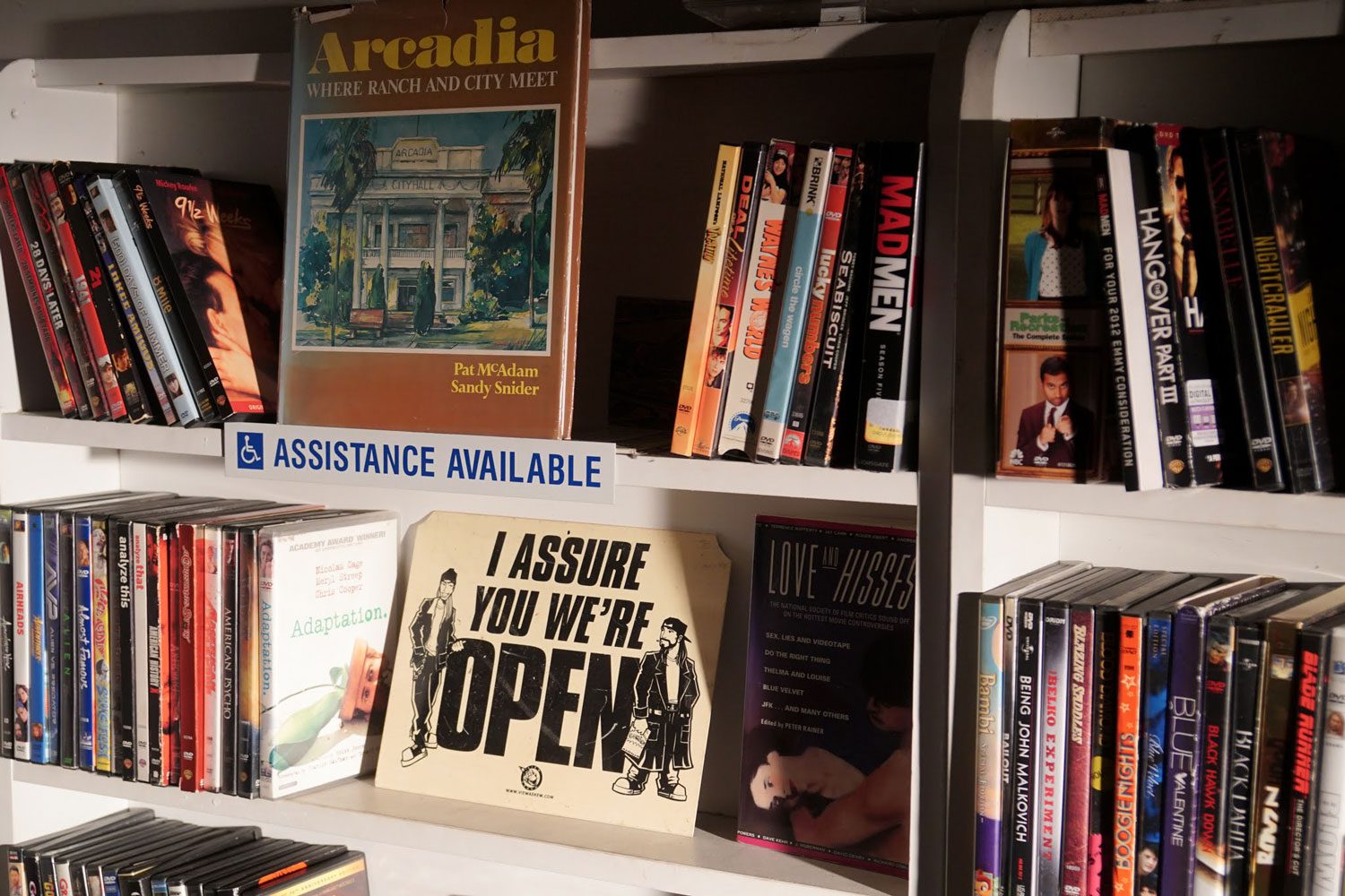decorated shelves of videos with "Assistance Available" and "I Assure you we're open" signs